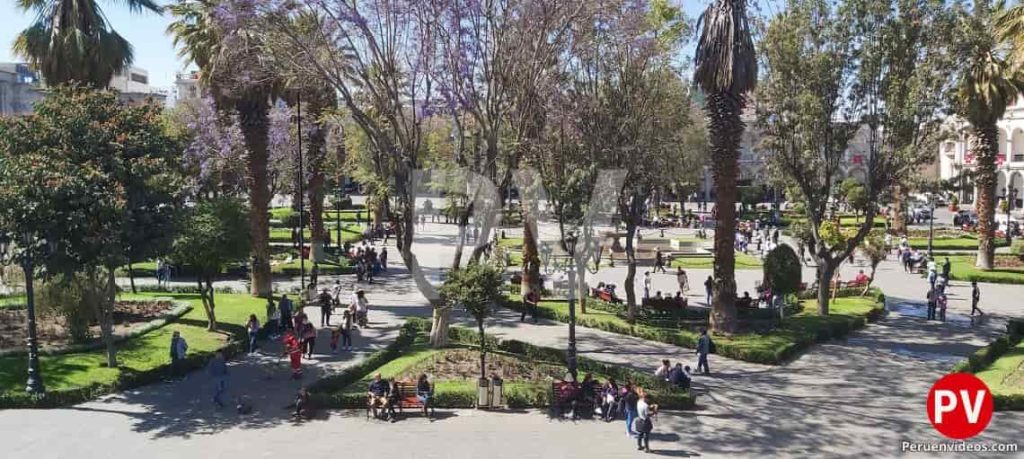 Jardines con árboles y palmeras rodeado de bancas y personas que cruzan sus veredas en la Plaza de Armas de Arequipa.