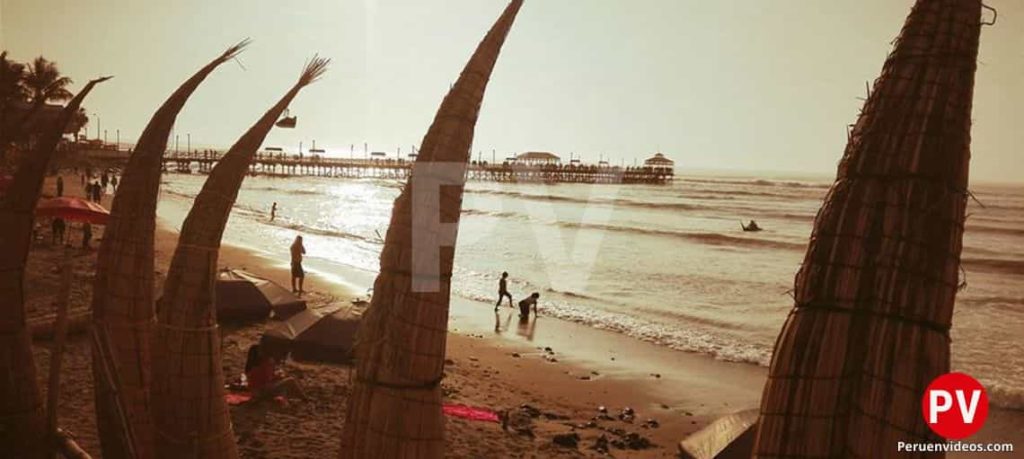 La tranquila playa y arena de Huanchaco con los caballitos de totora incrustados en la arena verticamente y algunas personas en las orillas mientras va cayendo el atardecer.