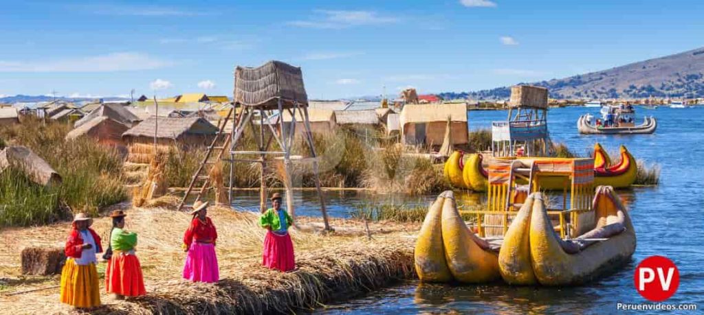 4 mujeres a orillas de una de las islas Los Uros frente a una totora. Atrás hay viviendas.